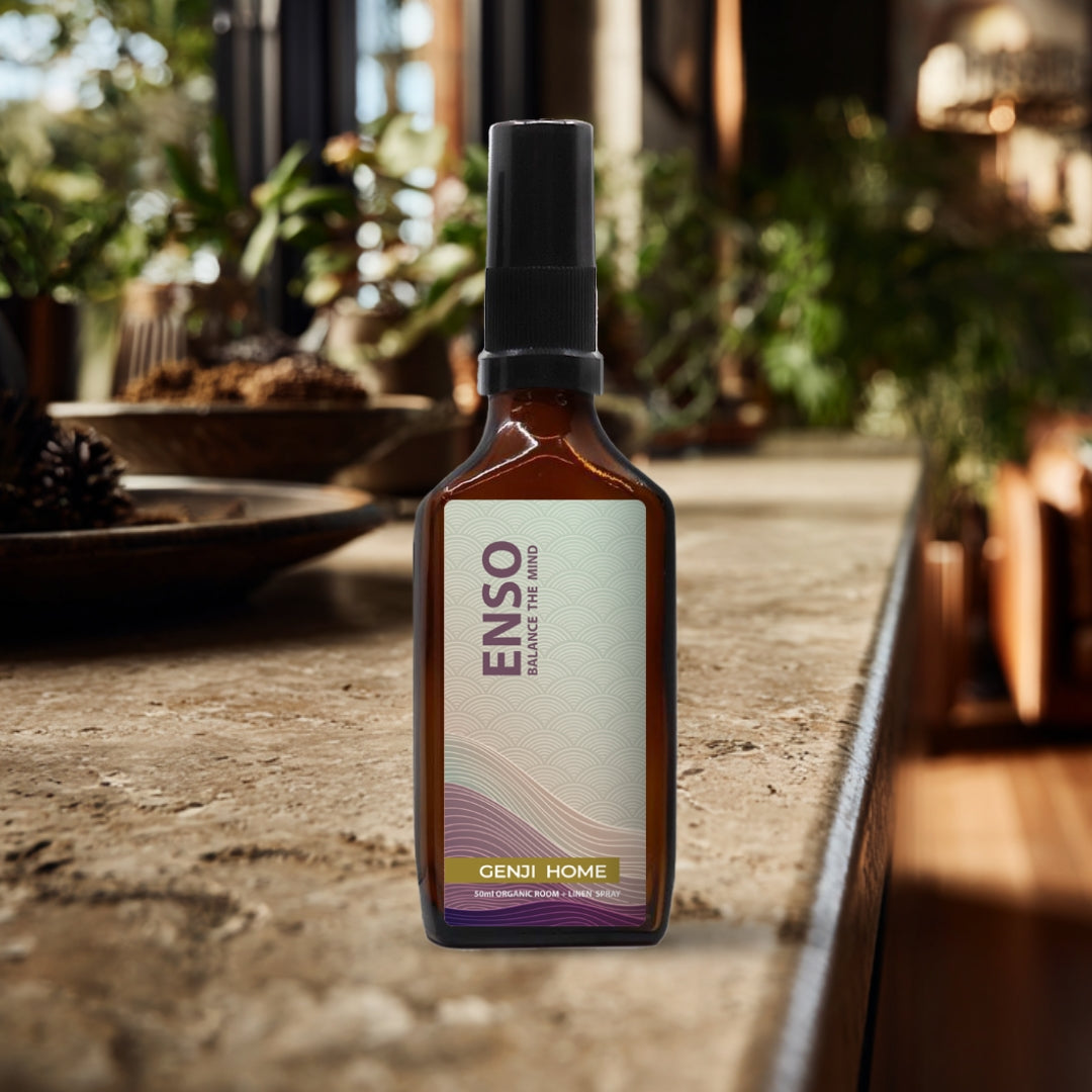 Enso room and linen spray by Genji Home in an amber bottle on a travertine surface with a blurred natural background of plants and decor.