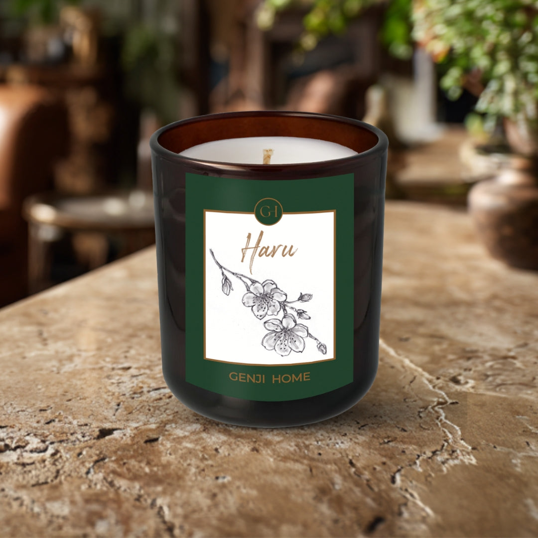 Haru Olive Wax Candle by Genji Home in an amber bottle on a travertine surface with a blurred natural background of plants and decor.