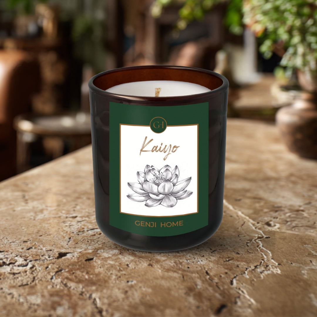 Kaiyo Olive Wax Candle by Genji Home in an amber bottle on a travertine surface with a blurred natural background of plants and decor.