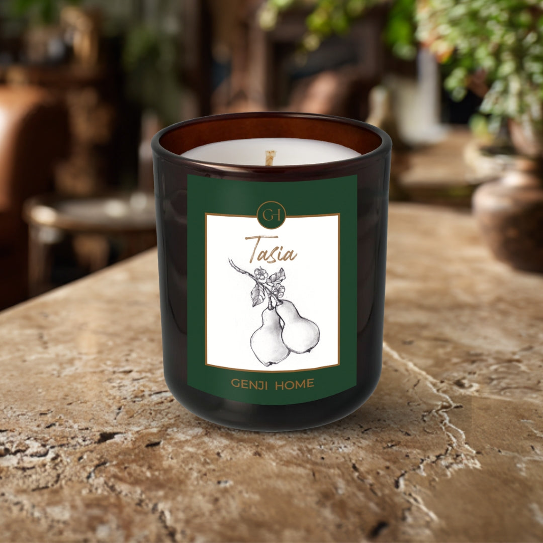 Tasia Olive Wax Candle by Genji Home in an amber bottle on a travertine surface with a blurred natural background of plants and decor.